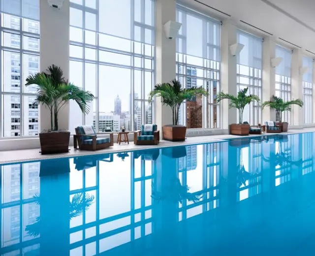 Indoor swimming pool with bright blue water, surrounded by large windows with city views, palm trees in planters, and lounge chairs.