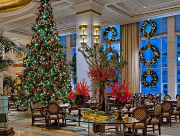 Elegant hotel lobby decorated for Christmas with a tall, illuminated tree, festive wreaths on windows, and plush seating areas.