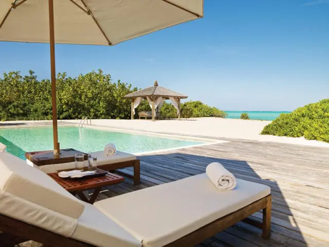 Wooden pool deck with lounge chairs, a swimming pool, and a gazebo overlooking a sandy beach and turquoise ocean.