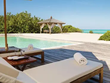 Wooden pool deck with lounge chairs, towels, and drinks next to a turquoise pool. A thatched-roof gazebo sits on the sandy beach beyond the pool, with lush green foliage and the ocean in the background.