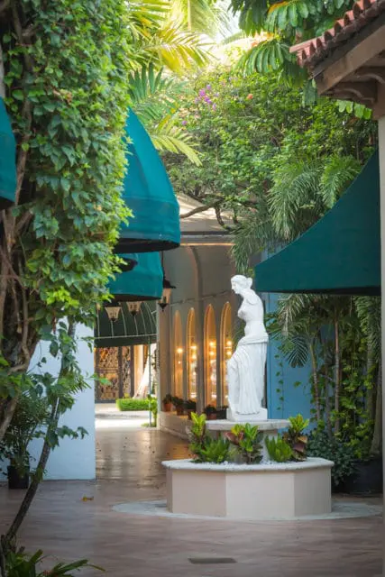 Courtyard with a white classical statue, lush green foliage, teal awnings, and arched windows with warm interior lighting.