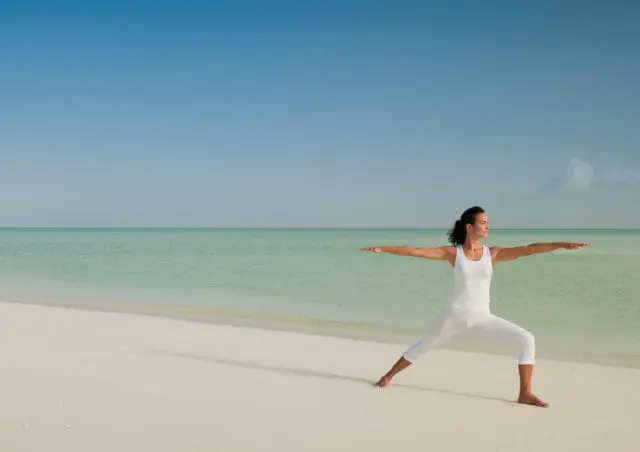 Woman in white tank top and capri pants practicing yoga warrior II pose on a sandy beach with turquoise water and clear blue sky.