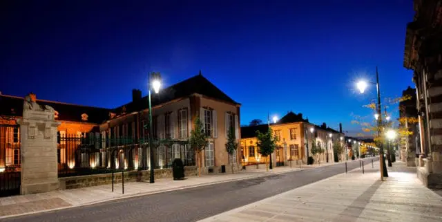 Row of illuminated buildings along a cobblestone street at dusk, with ornate lampposts and trees lining the sidewalk.