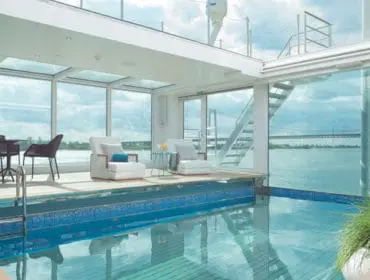 Indoor pool area with white lounge chairs, a small table, and a dining set, surrounded by large windows overlooking a body of water and a bridge.