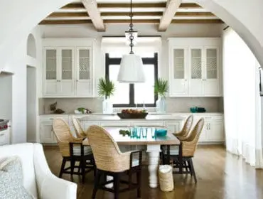 White kitchen with beamed ceiling, white cabinetry featuring lattice-style doors, and a dining area with woven chairs around a light wood table.