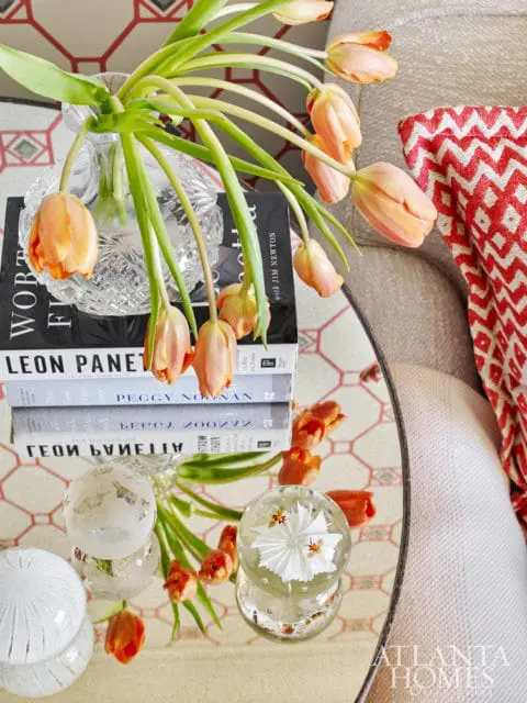 Stack of books on a mirrored surface with a vase of peach tulips and decorative glass orbs. A red and white patterned throw is draped over a chair.