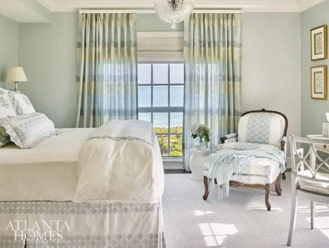 Bedroom with light blue walls, striped curtains, a bed with white linens and patterned skirt, an armchair with geometric pattern, and a desk with a chair.