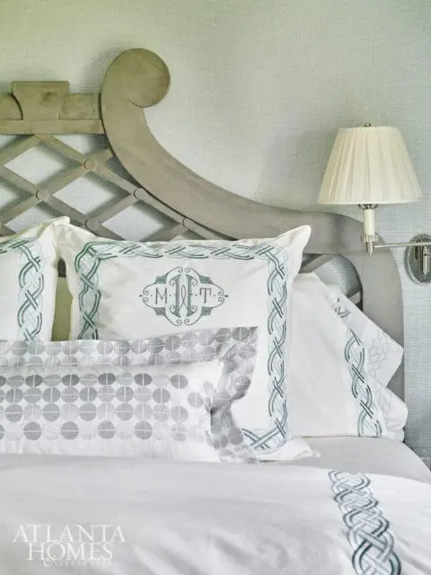 Close-up of a bed with white linens, green embroidered Celtic knot trim, and a central pillow with a monogrammed crest. A grey wooden headboard with lattice detail is visible.