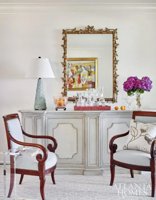Light blue upholstered chairs flank a distressed grey sideboard topped with a gold ornate mirror, colorful painting, glassware, and a lamp.