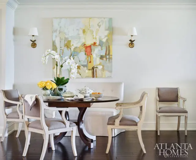 Dining room with round wooden table, upholstered chairs, banquette seating, abstract art, and wall sconces.