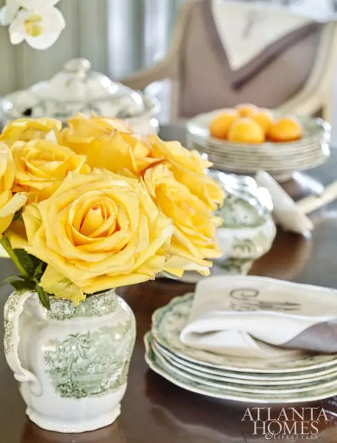 A bouquet of yellow roses in a green and white pitcher sits on a dark wood table next to stacked plates and a napkin.