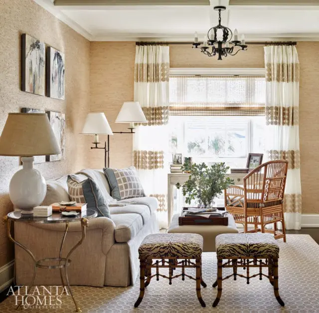 Living room with grasscloth wallpaper, a light gray sofa with patterned pillows, a glass-top side table, two animal-print ottomans, and a desk with a rattan chair by the window.