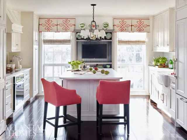 White kitchen with a pink island, two coral upholstered bar stools, dark wood floors, and a black chandelier.