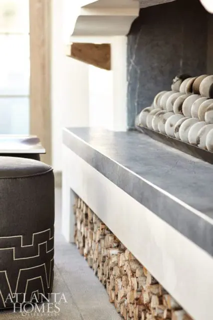 Dark gray upholstered ottoman with geometric pattern next to a fireplace with a gray stone hearth and stacked firewood below