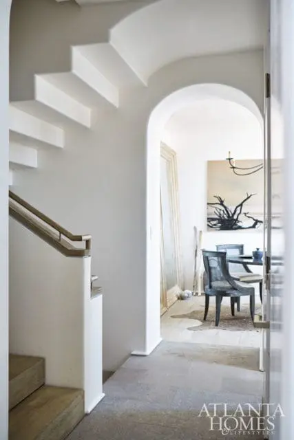 White plaster staircase with wooden handrail leading to an arched doorway revealing a dining area with a dark wood table, chairs, and abstract art