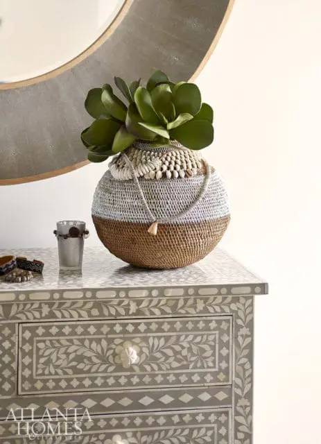 Woven basket with shells and a necklace holding faux succulents sits on a bone inlay dresser with floral patterns.