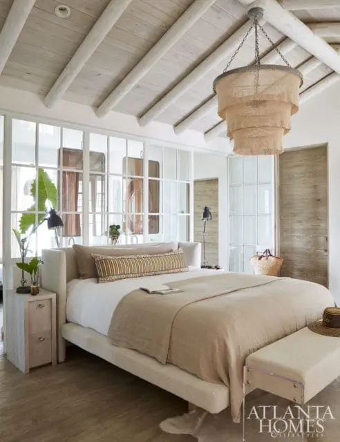 Bedroom with a light-colored upholstered bed, linen bedding, and a tiered rattan chandelier. A glass-paneled wall separates the room from a closet area.