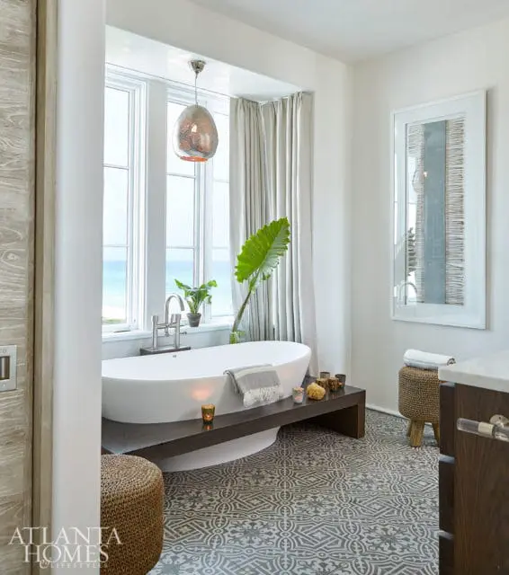 Modern bathroom with freestanding tub, patterned tile floor, large windows overlooking the ocean, and a decorative pendant light.