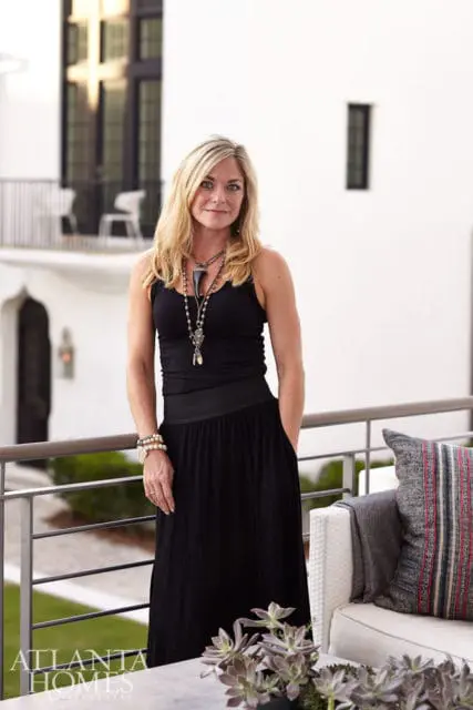 Woman with blonde hair wearing a black tank top and long black skirt, standing on a balcony with a white building facade behind her.