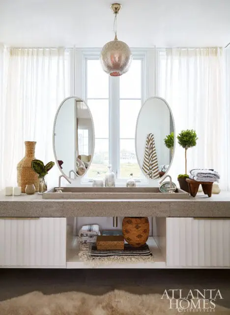 Modern bathroom with a double concrete vanity, oval mirrors, fluted white cabinets, and a large window with sheer curtains.