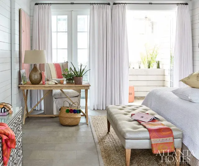 Bedroom with white shiplap walls, light wood desk with lamp and plants, tufted bench with colorful throw, and white curtains opening to a balcony.