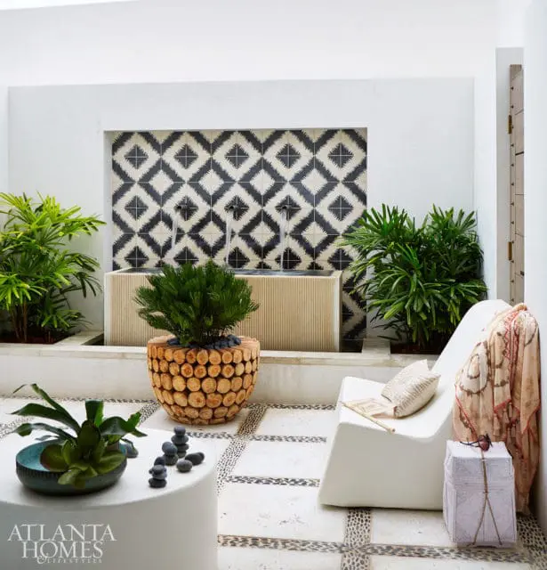 Courtyard with a black and white geometric tile fountain, a white lounge chair draped with a patterned scarf, and a round wooden planter filled with a small tree.