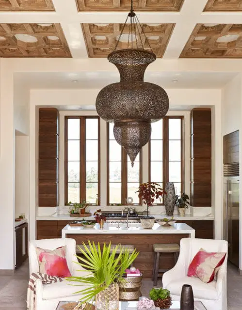 Living area with white armchairs and a kitchen island in the background, featuring a large ornate metal chandelier and a coffered wood ceiling.