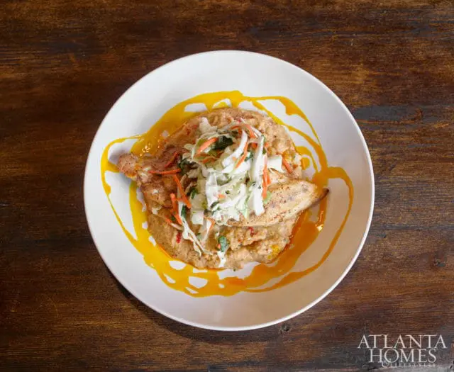 White bowl of pan-seared fish with grits and coleslaw, drizzled with orange sauce, on a dark wood table. Atlanta Homes & Lifestyles logo in the bottom right corner.