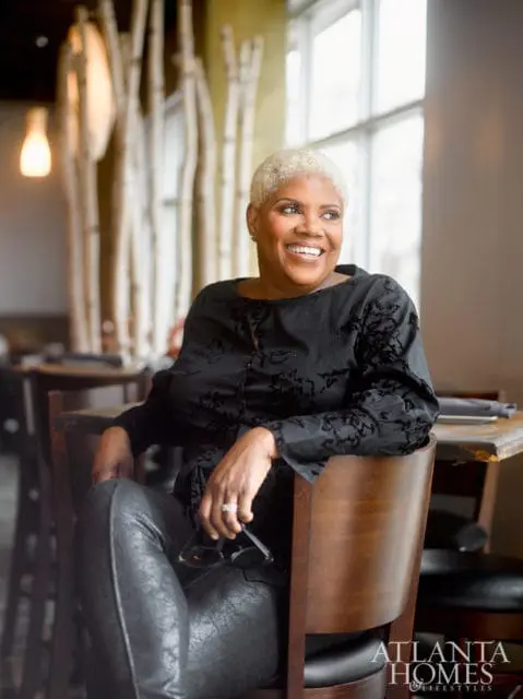 Woman with short blonde hair wearing a black patterned blouse and black pants, sitting in a wooden chair and holding glasses.