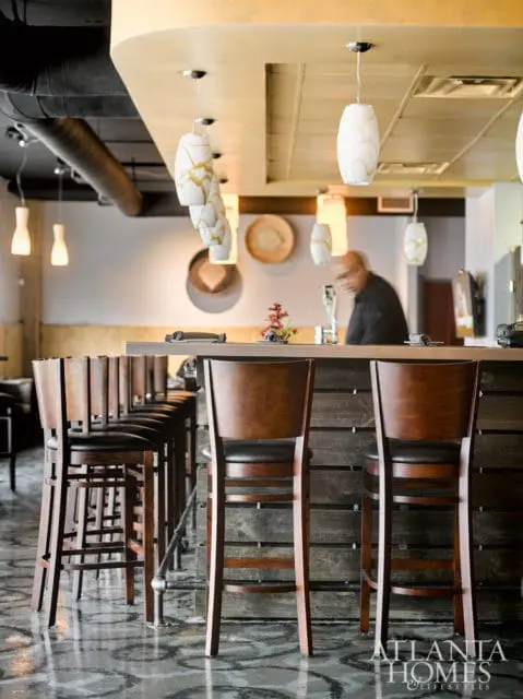 Bar interior with rows of dark wood bar stools with black leather seats in front of a wooden slatted bar. Pendant lights hang from a gold ceiling.