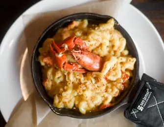 Close-up of creamy macaroni and cheese topped with lobster meat and claws, served in a black cast iron skillet on a white plate.