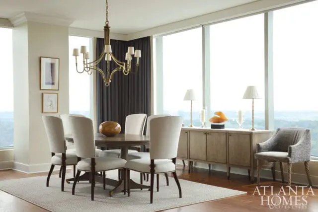 Dining room with a round wooden table and upholstered chairs, a brass chandelier, and a long buffet with lamps and decorative objects.