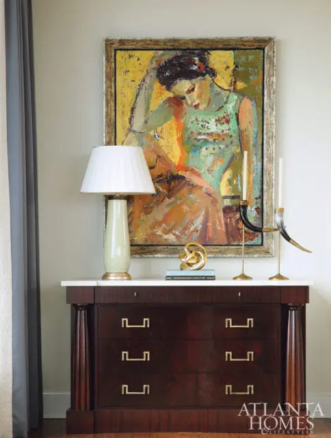 Dark wood dresser with marble top, brass hardware, and a table lamp, abstract sculpture, and artwork above.