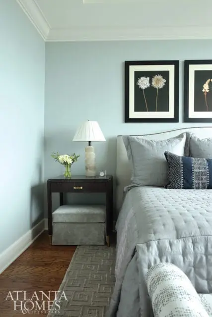 Light blue bedroom with a gray quilted bed, dark wood nightstand with a stacked stone lamp, and framed botanical prints on the wall.