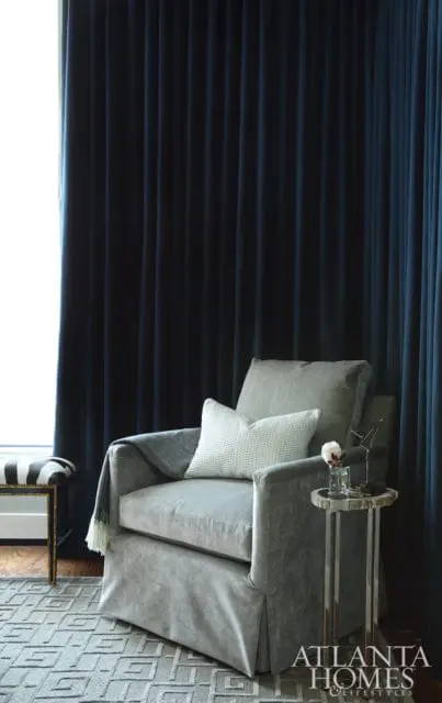 Gray slipcovered armchair with a textured white pillow and a gray throw blanket, next to a small metallic side table with a glass vase and a flower. Dark blue curtains frame the scene.