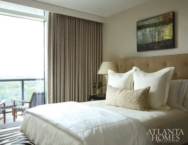 Bedroom with a tufted headboard, white quilted bedding, and a patterned accent pillow. A landscape painting hangs above the bed, and a large window with patterned curtains reveals an outdoor balcony.