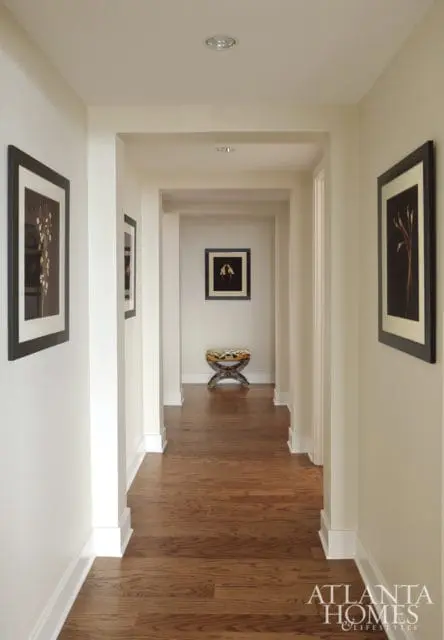 Hallway with cream walls, dark wood floors, and framed botanical prints. A decorative stool sits at the end of the hall.