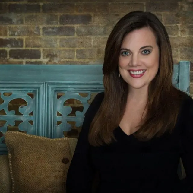 Jessie La Falce smiles, wearing a black V-neck shirt, seated in front of a teal distressed headboard and a brick wall.