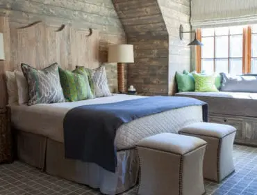 Rustic bedroom with a wooden plank headboard and walls, a bed with a blue throw and patterned pillows, and a window seat with green cushions.