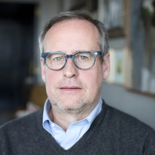 Man with graying hair and blue-framed glasses wearing a light blue collared shirt and dark gray V-neck sweater.