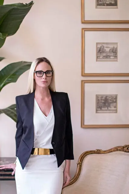 Woman with blonde hair and glasses wearing a black blazer over a white top and skirt with a gold belt, standing next to a large plant and an ornate chair.