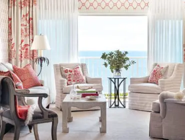 Living room with red and white toile curtains, striped armchairs, and a view of the ocean through the window.