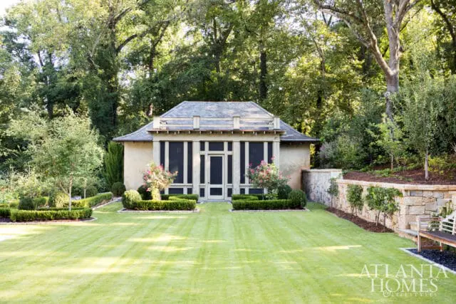 A stucco building with a screened-in porch and a gray shingle roof sits behind a manicured lawn and formal gardens. A stone retaining wall with a wooden bench is to the right.