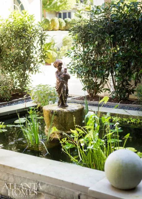 Bronze statue of a child holding a pitcher in a garden pond with water lilies and lush greenery, surrounded by manicured bushes and a stone pathway.