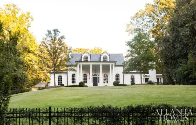 White Georgian-style mansion with a central portico supported by columns, arched windows, and dormers, set on a manicured lawn with mature trees and a black wrought-iron fence.