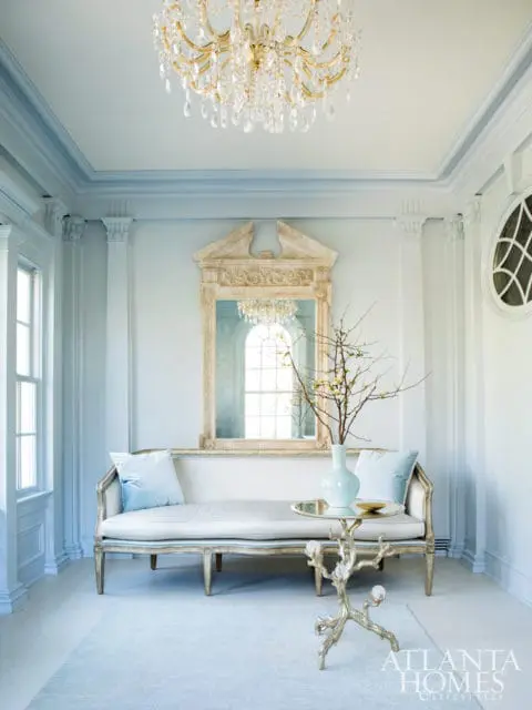 Elegant entryway with a light blue and white color palette, featuring a gilded crystal chandelier, a carved mirror, a pale blue settee with velvet pillows, and a decorative side table with a branch motif.