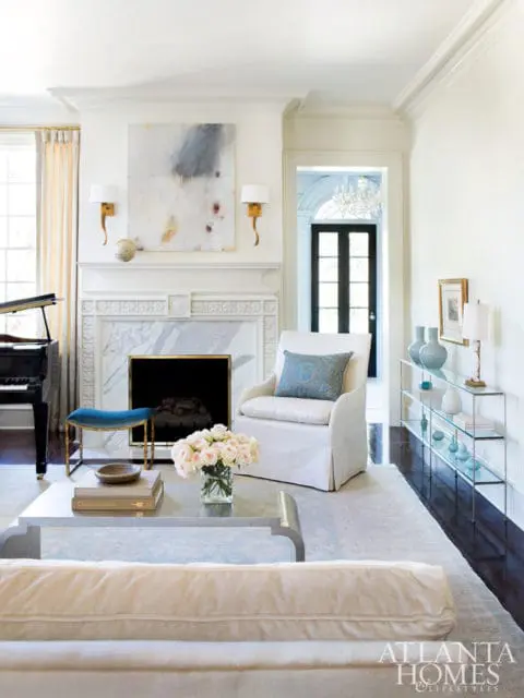 Living room with a white slipcovered armchair, marble fireplace, abstract art, and a grand piano. A glass console table with blue vases is visible in the background.
