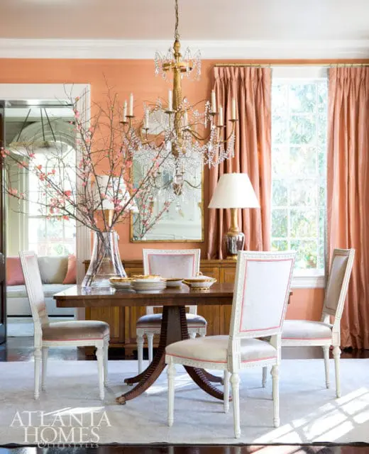 Dining room with peach walls, a wooden dining table set with plates, and upholstered chairs. A crystal chandelier hangs above.