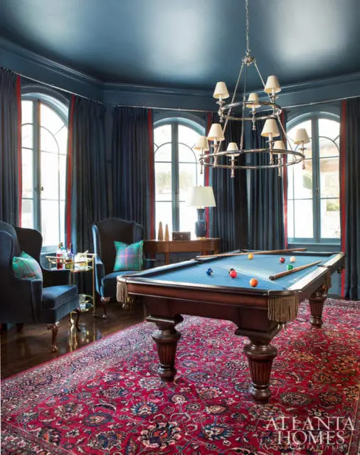 Dark blue game room with a pool table, plush armchairs, a large Persian rug, and arched windows with dark blue curtains.