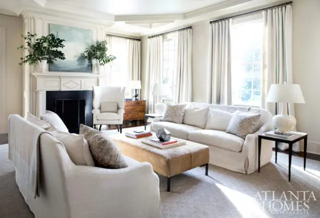 Living room with white slipcovered sofas, wingback chair, fireplace with abstract art, and a textured ottoman coffee table.